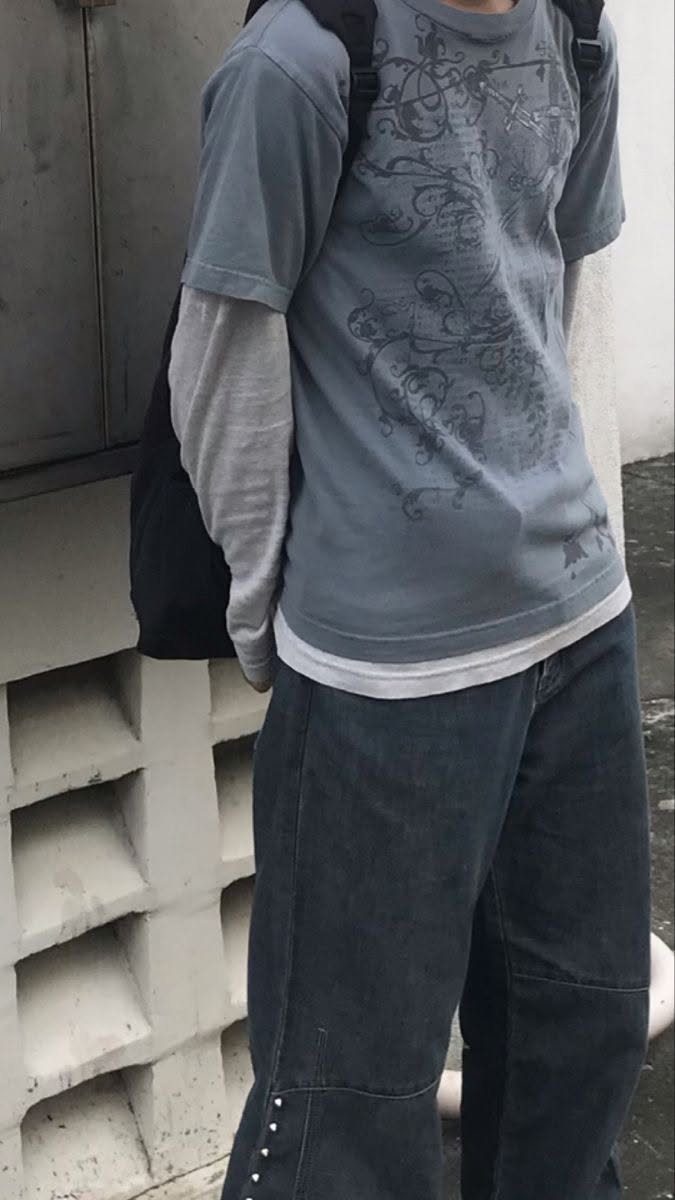 A person wearing a faded blue graphic t-shirt layered over a long-sleeved grey shirt and dark pants.
