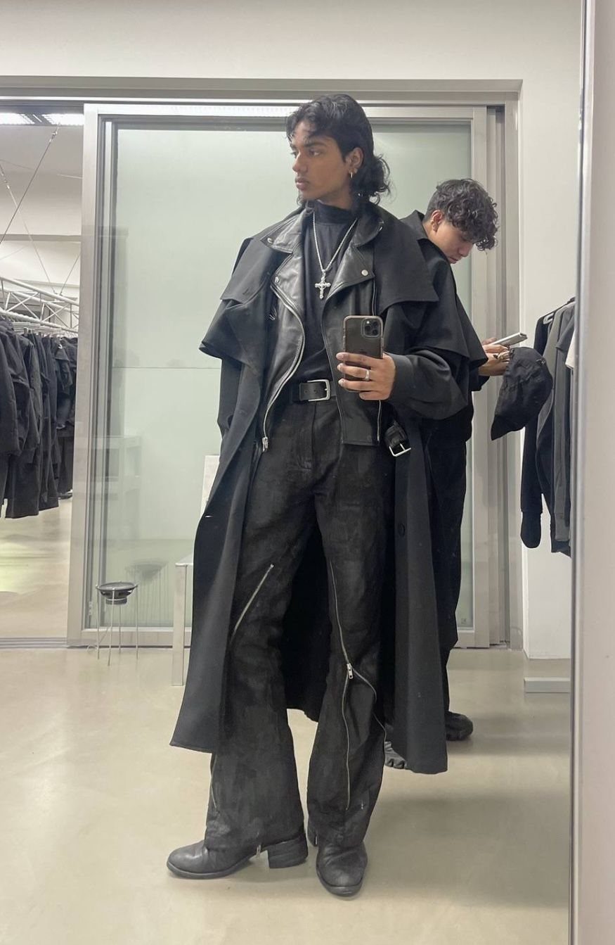 A person wearing a long black gothic coat, black trousers, and a silver cross necklace.