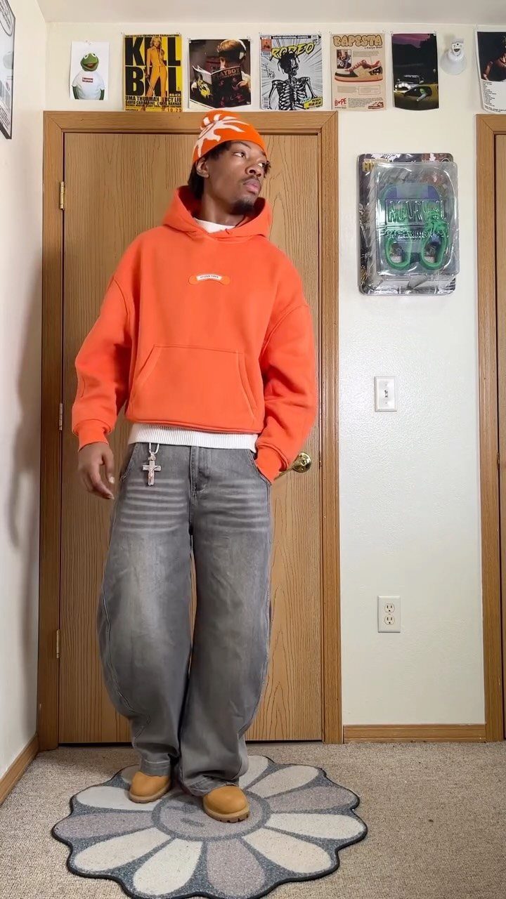 A man posing in an orange hoodie, orange beanie, and baggy grey jeans indoors.