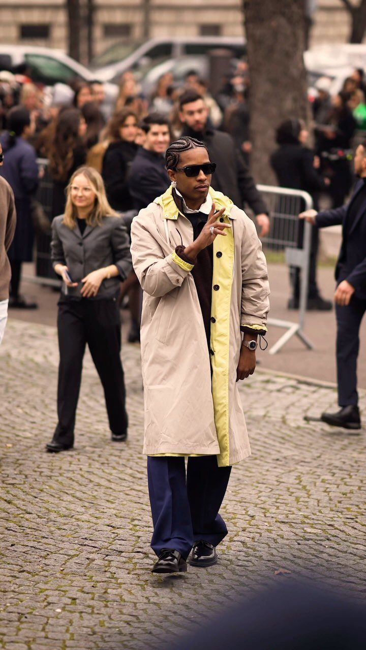 A man in a long beige and yellow trench coat walking through a crowded city street.