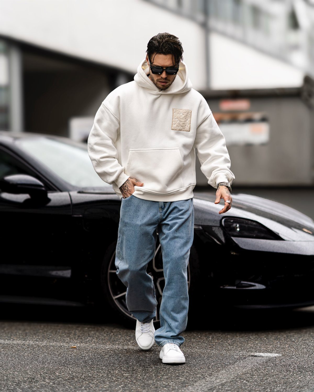 Man in a white hoodie and blue jeans walking past a black car.