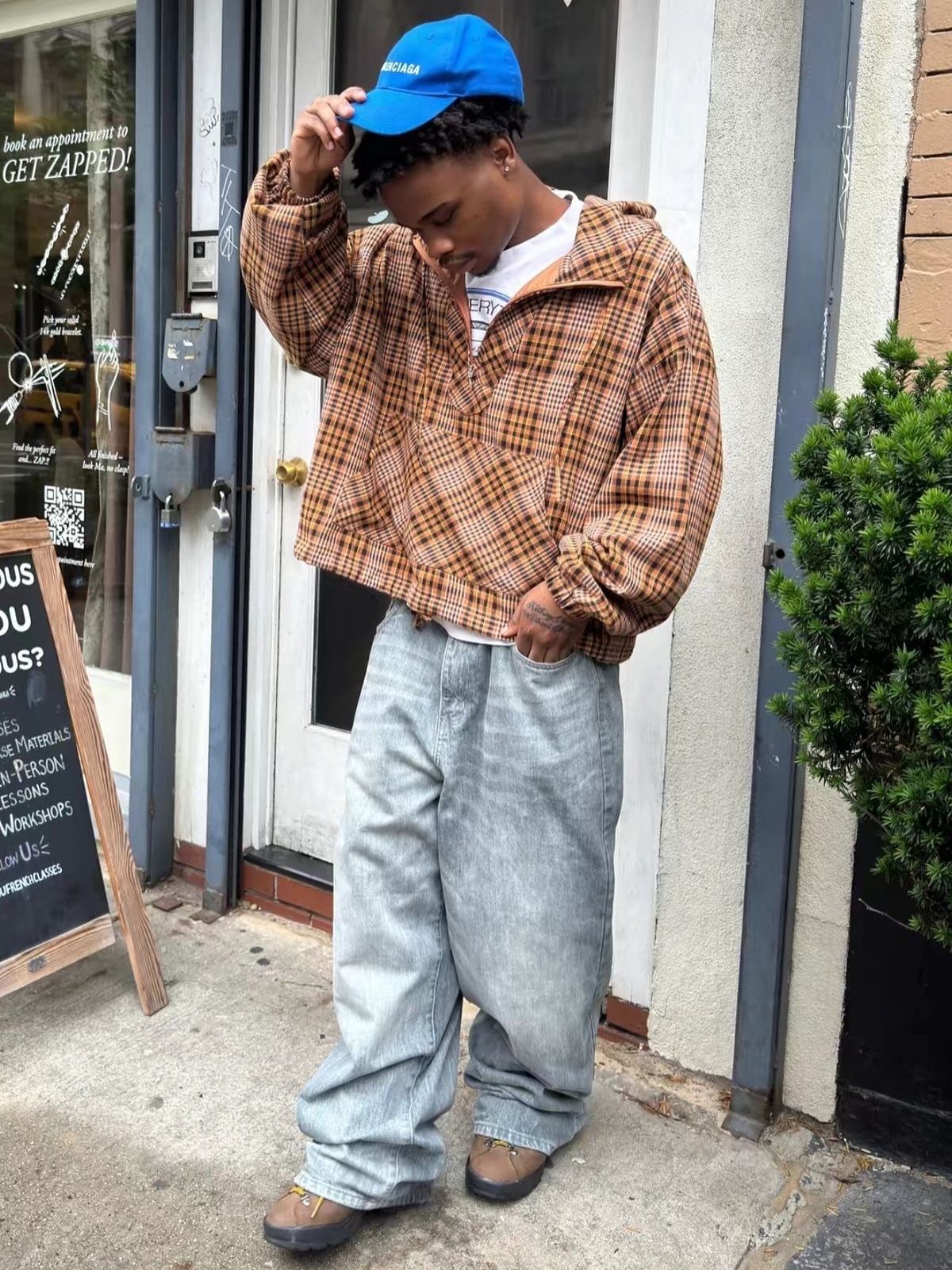 A young man in a plaid pullover and baggy jeans posing on a city sidewalk.