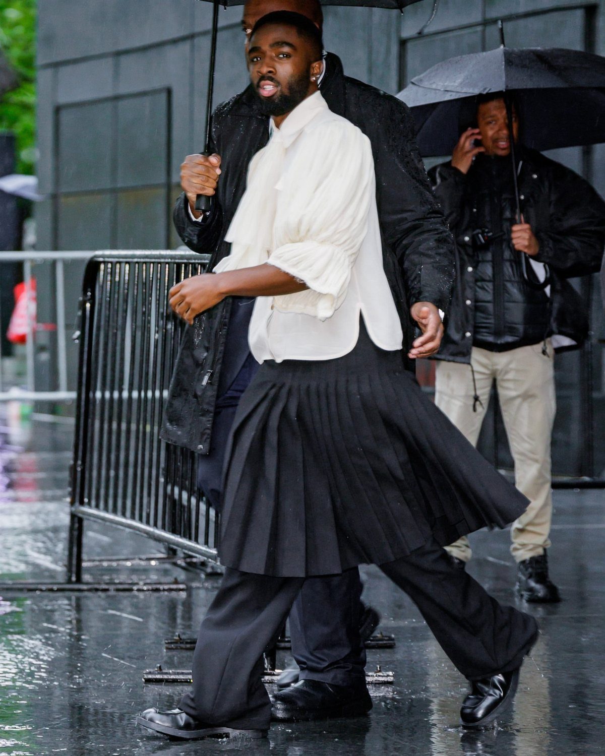 Man in a white ruffled blouse and black pleated skirt walking through the rain.