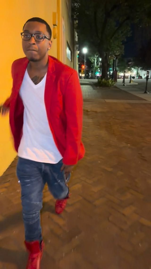A man in a red blazer, white t-shirt, and blue jeans walking outdoors at night.