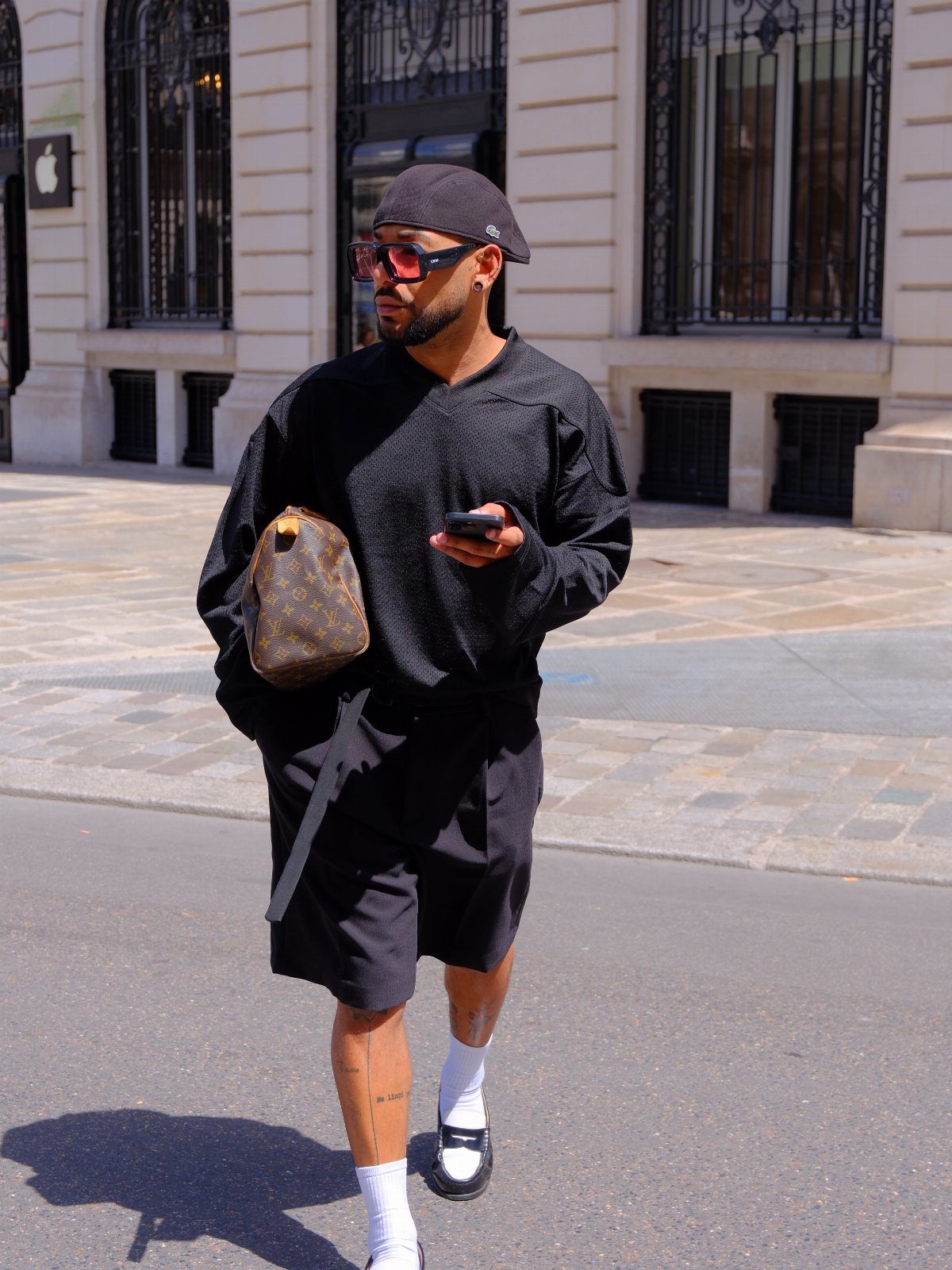Man in black streetwear walking down a city street wearing pink sunglasses and carrying a brown bag.