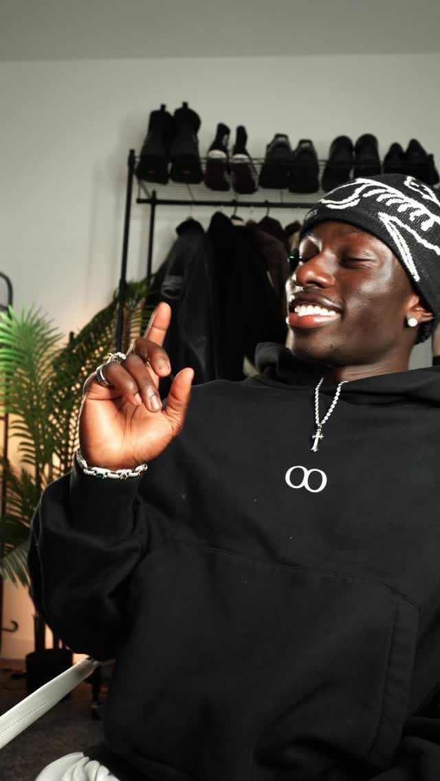 A smiling man wearing a patterned beanie, cross necklace, silver bracelet, and rings.