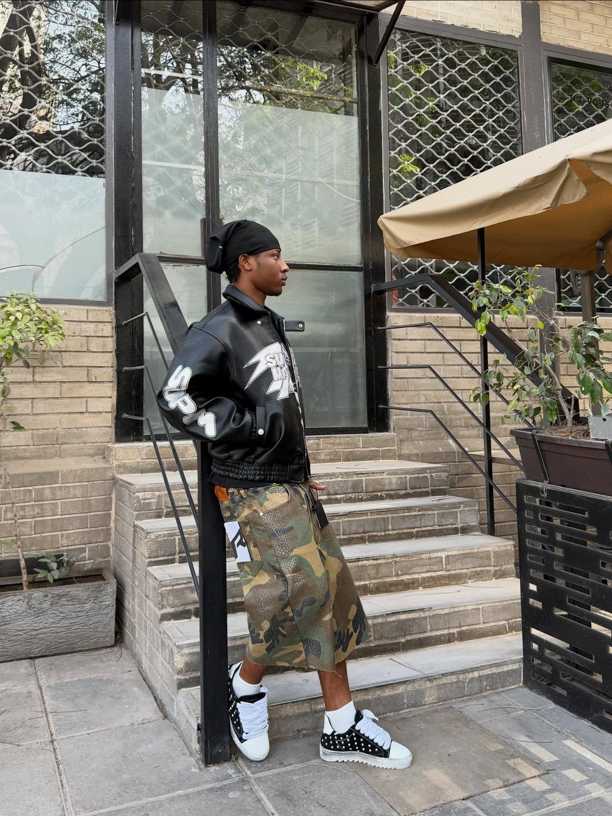 A man in a graphic leather jacket and camouflage shorts posing on outdoor stone steps.