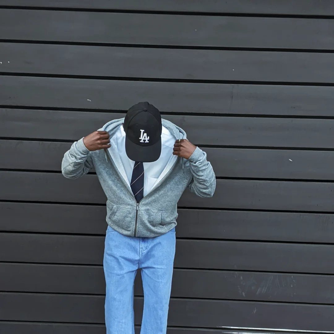 Person in a grey hoodie, tie, and blue jeans standing against a dark slatted wall.