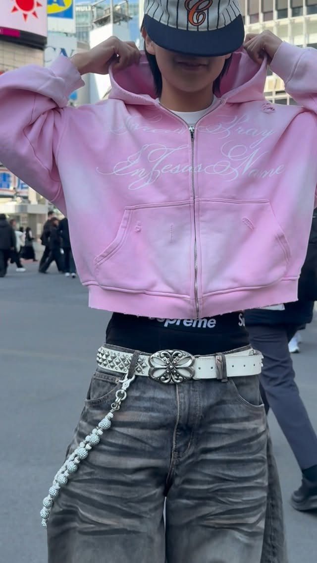Person wearing a pink cropped hoodie, distressed jeans, and a studded white belt with a silver chain.