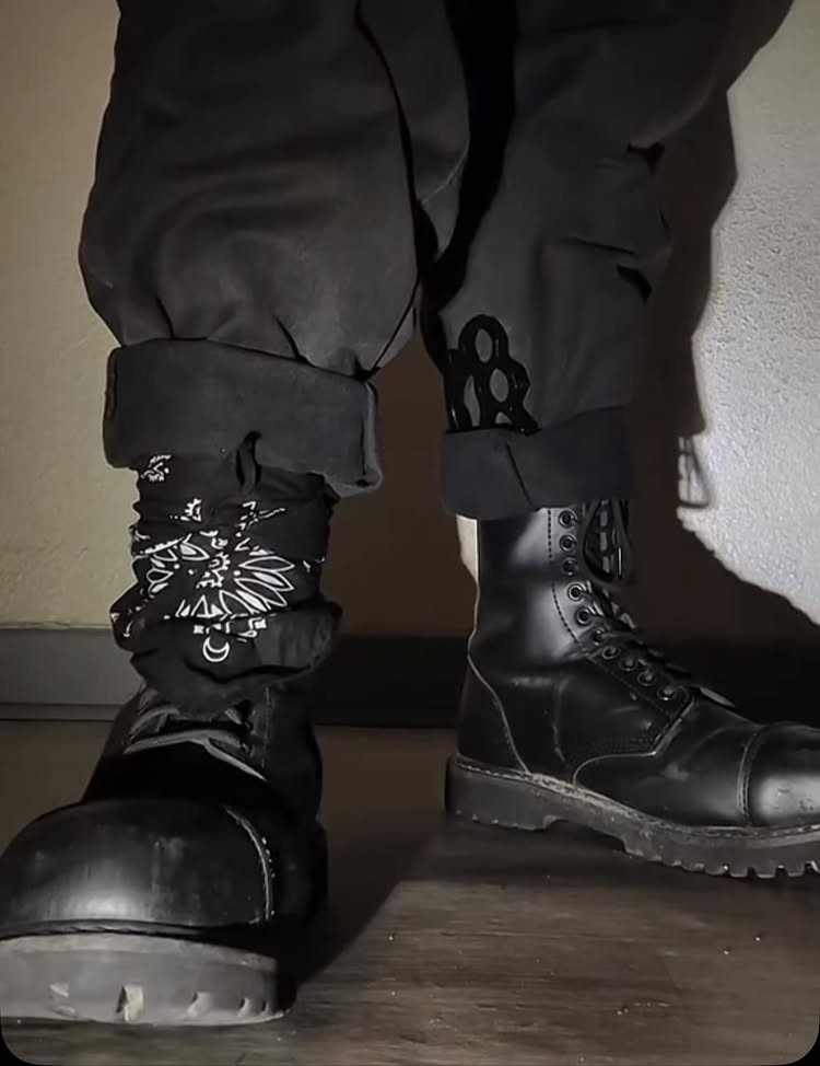 Black combat boots worn with patterned socks and cuffed black trousers on a wooden floor.