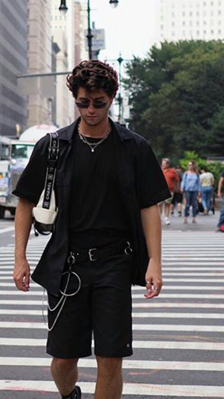 A young man in all black summer clothing wearing dark sunglasses while walking across a city street.