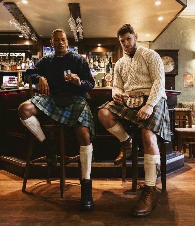 Two men sitting on bar stools wearing traditional tartan kilts and long socks in a pub.