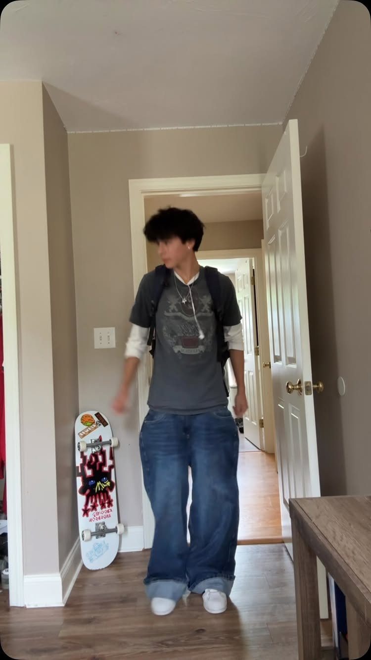 A person in baggy jeans and a graphic tee walks through a doorway with a skateboard nearby.
