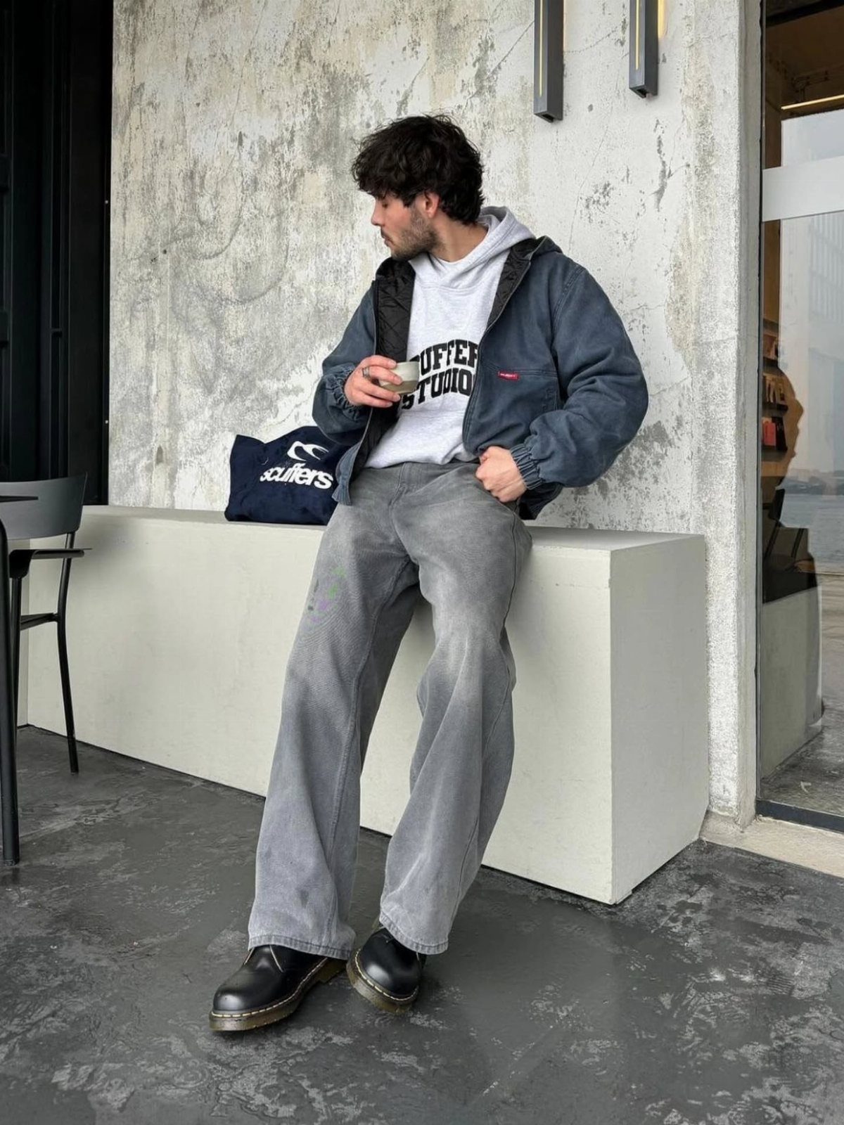 Man in baggy grey jeans and a blue bomber jacket leaning against a white wall.