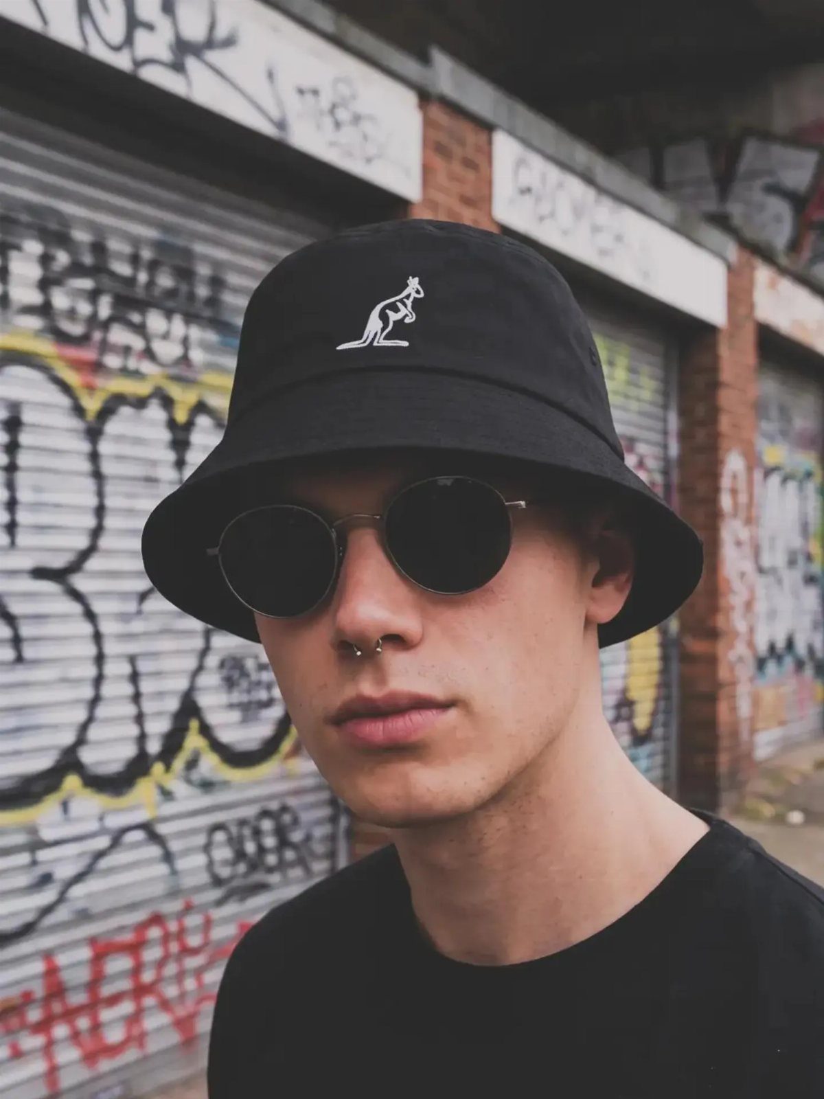 Young man wearing a black bucket hat and round sunglasses against a graffiti background.