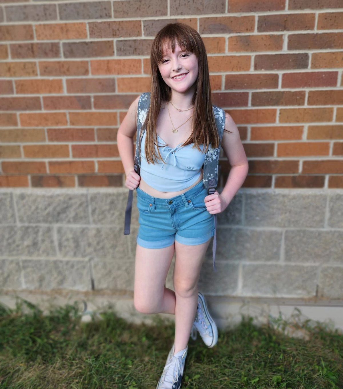 Smiling teenage girl wearing a blue halter top, denim shorts, and high-top sneakers against a brick wall.