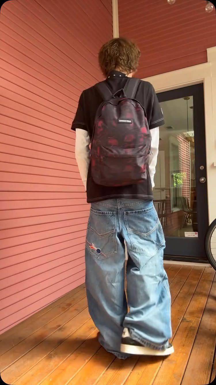 Person wearing baggy blue jeans and a dark backpack standing on a wooden porch.