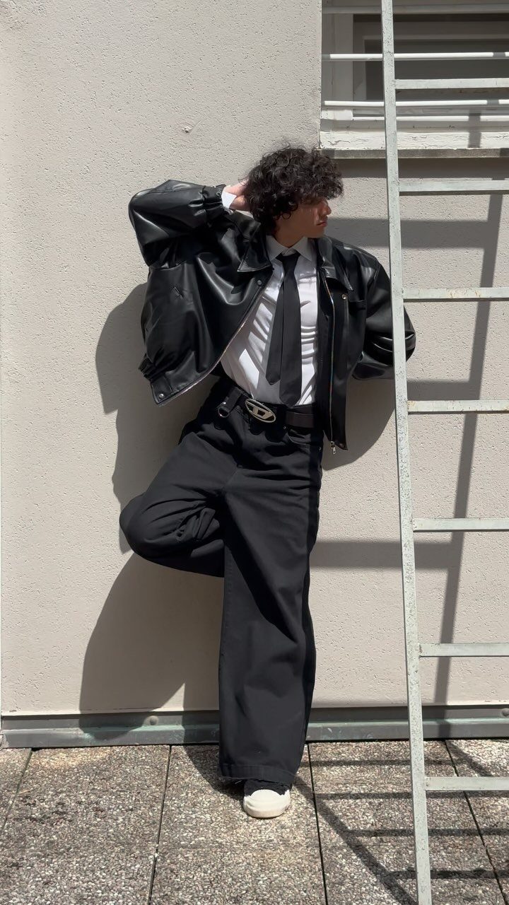 A person wearing an oversized black leather jacket, white shirt, and black tie posing against a wall.