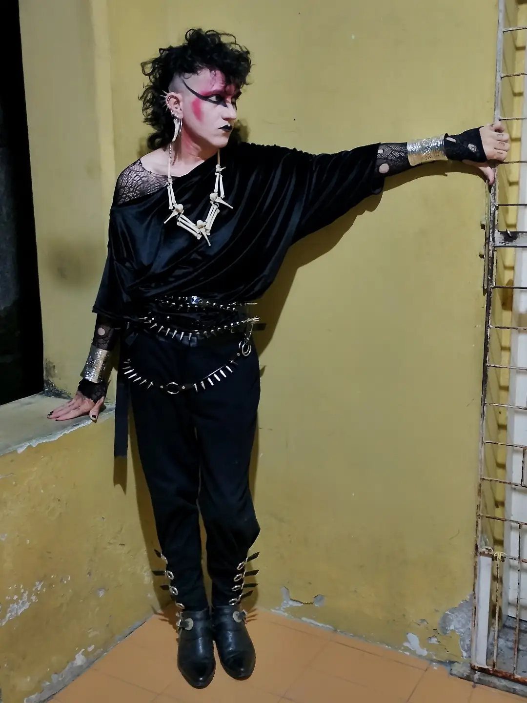 Person in deathrock fashion with teased hair, dramatic makeup, and studded black clothing posing against a wall.
