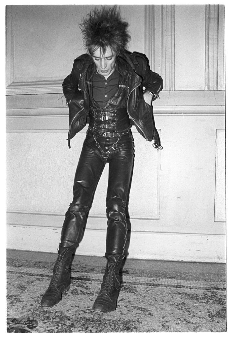 A person with teased black hair wearing leather clothing and boots posing against a wall.