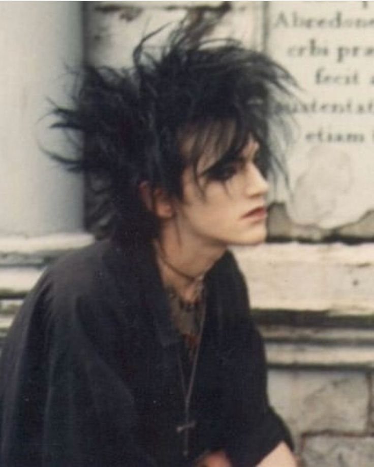 A person with spiky black hair and dark makeup wearing a black shirt and cross necklace.