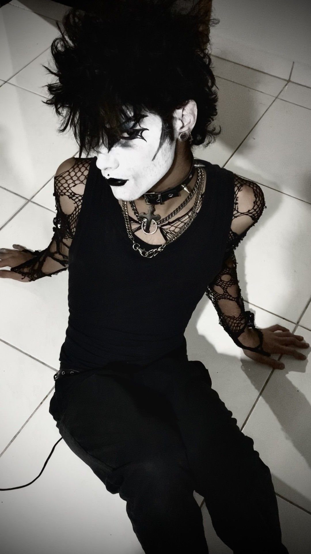 A person with pale makeup and black hair wearing fishnet sleeves and silver necklaces.