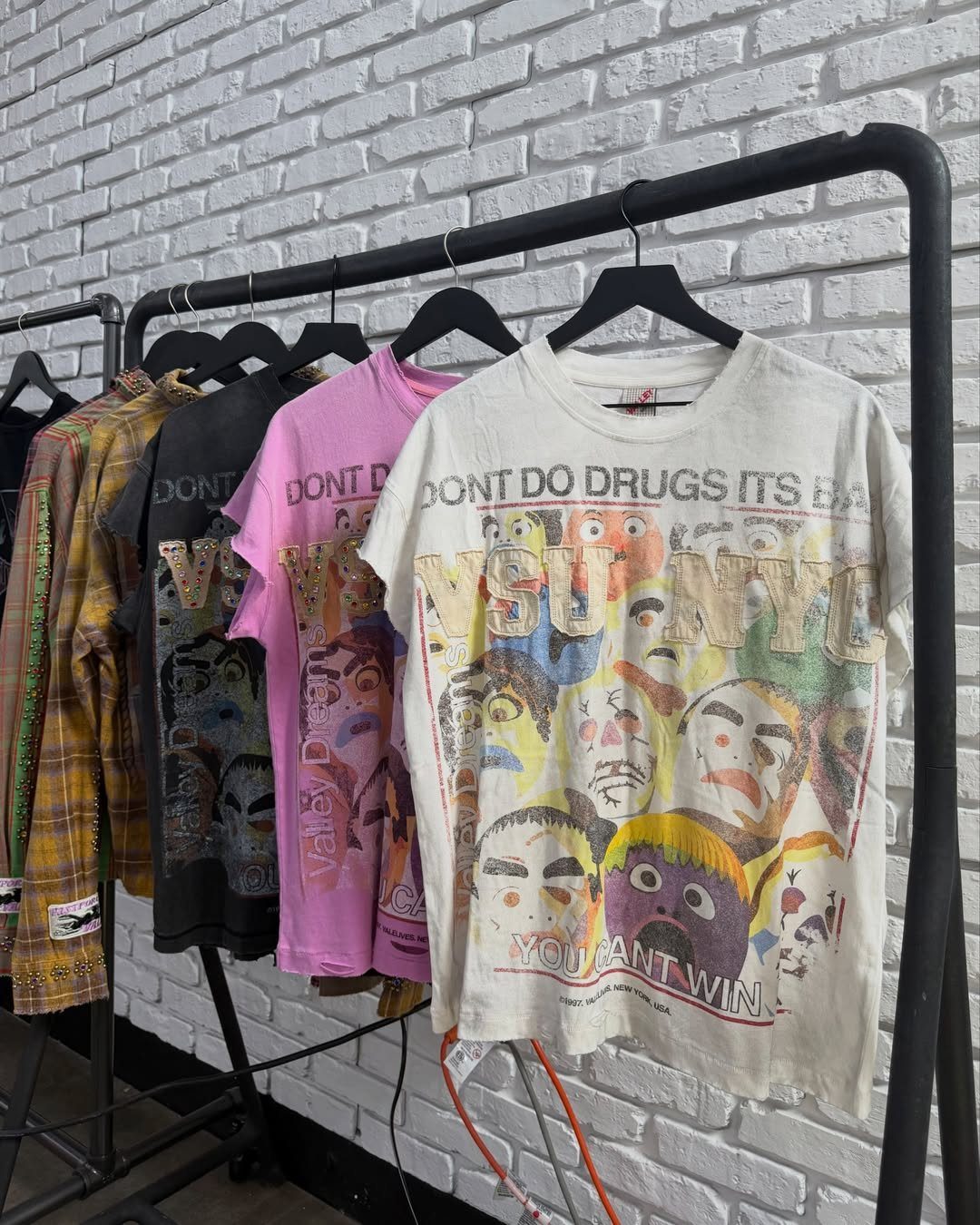 Graphic t-shirts with vintage character prints hanging on a black clothing rack against a brick wall.