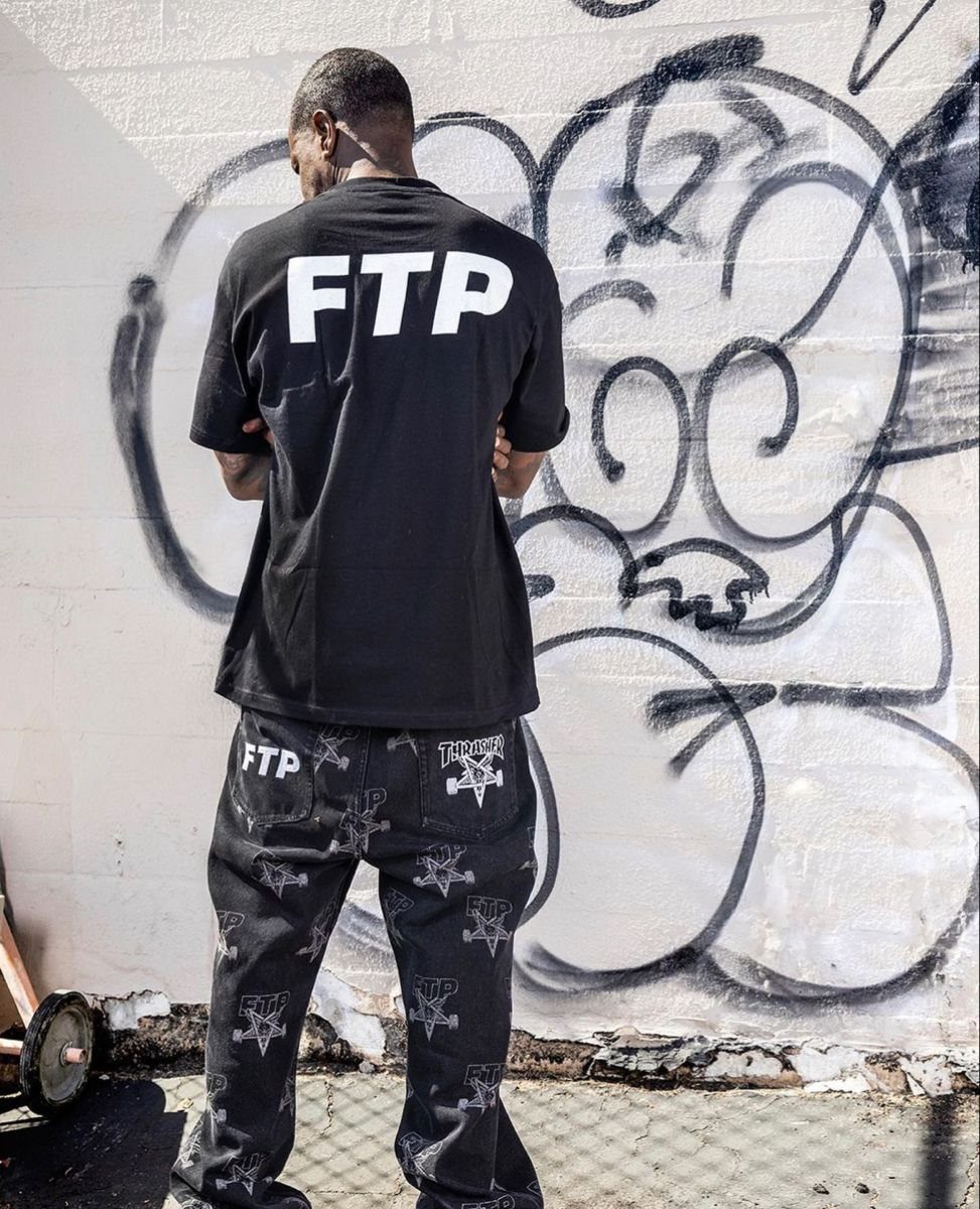 A man wearing a black FTP t-shirt and patterned pants stands before a graffiti wall.