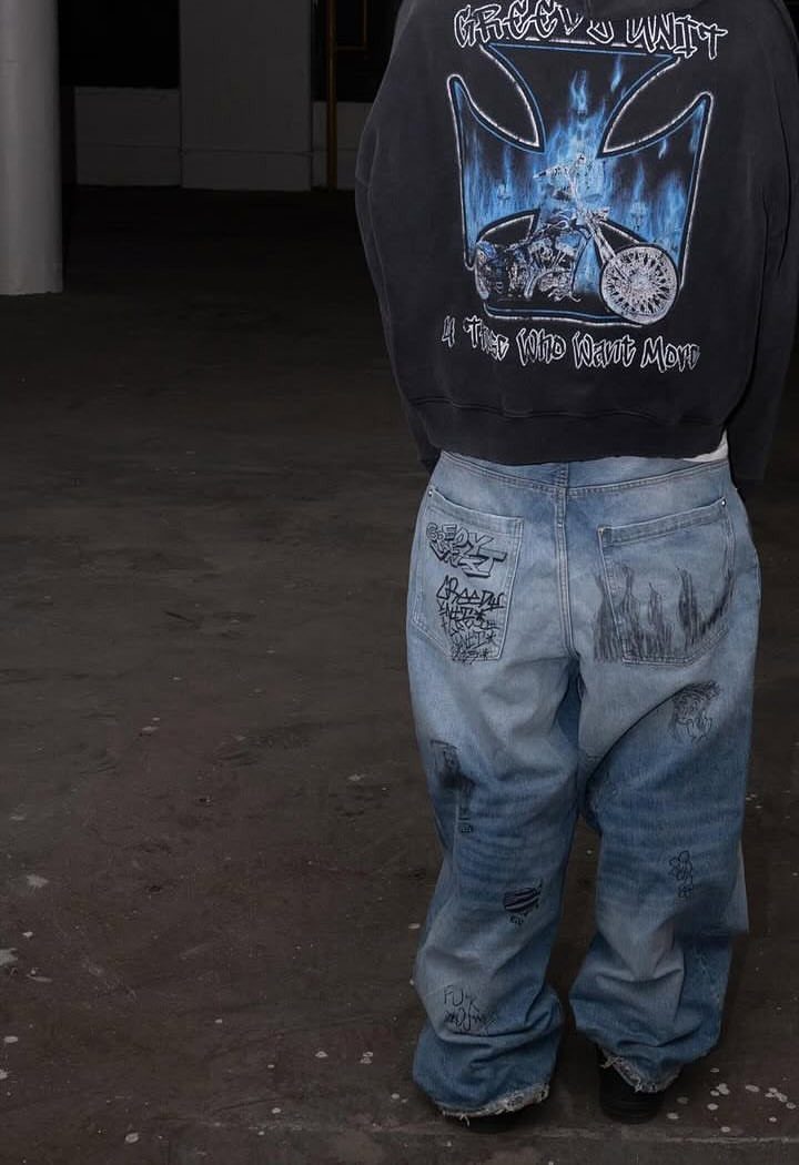 Person wearing a dark graphic sweatshirt and baggy printed denim jeans standing on concrete.