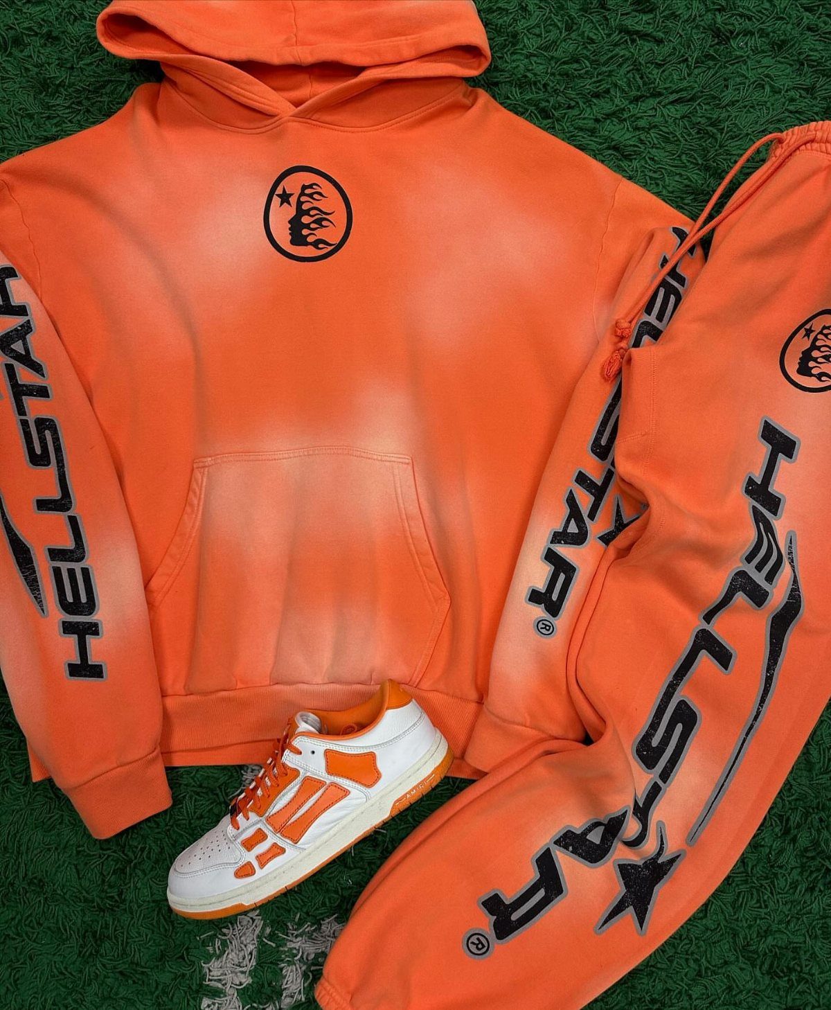 Orange Hellstar hoodie and sweatpants set with a white and orange sneaker on green grass.