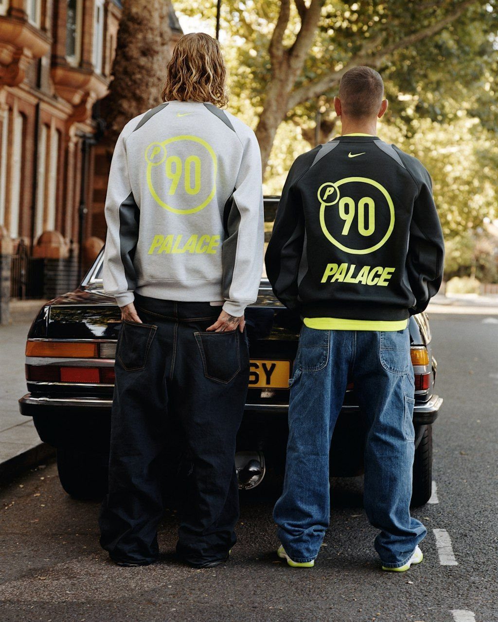 Two men wearing Palace branded streetwear jackets and baggy jeans standing by a car.