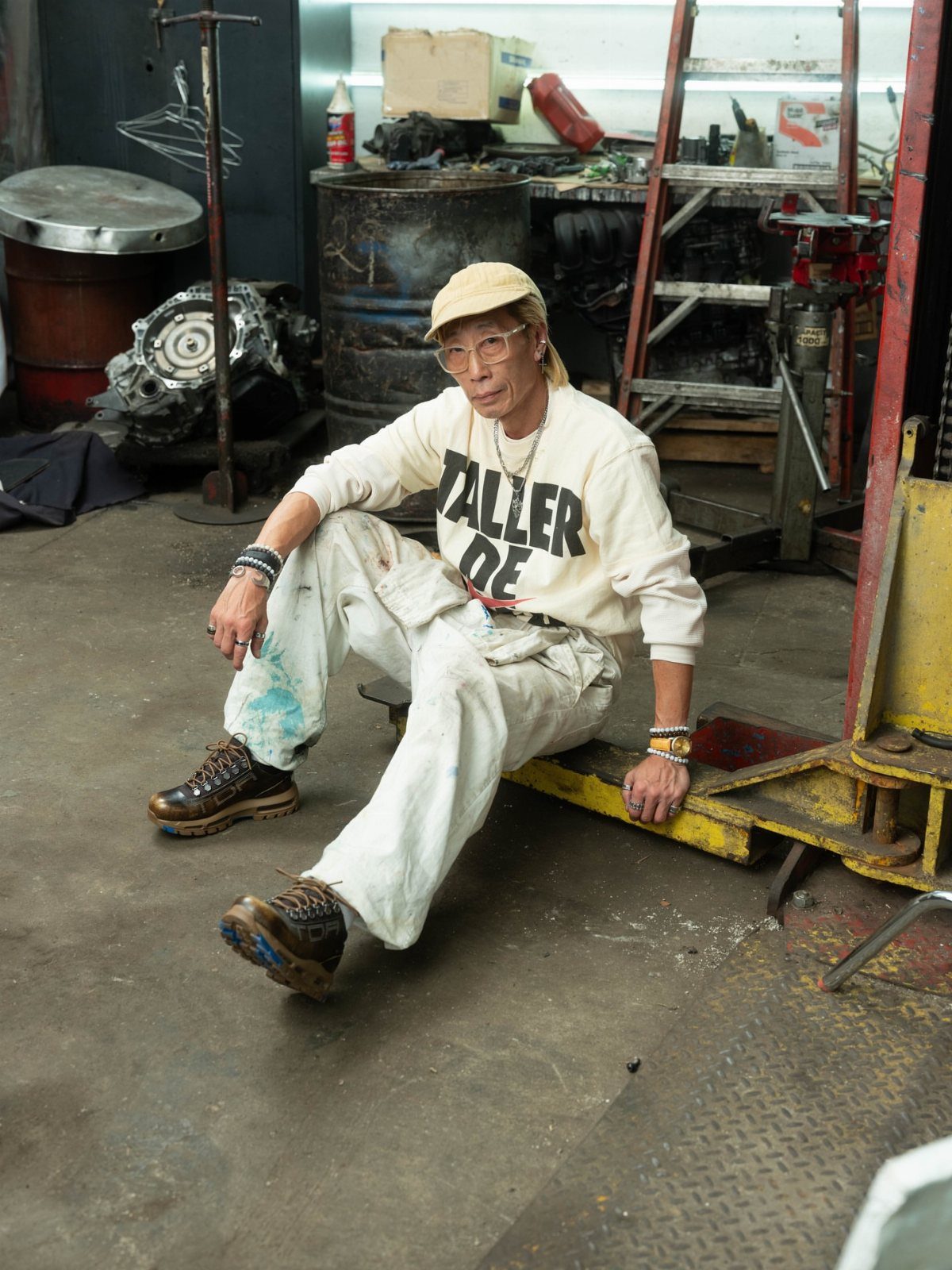 Man in cream sweatshirt and white pants sitting in a garage surrounded by automotive parts.
