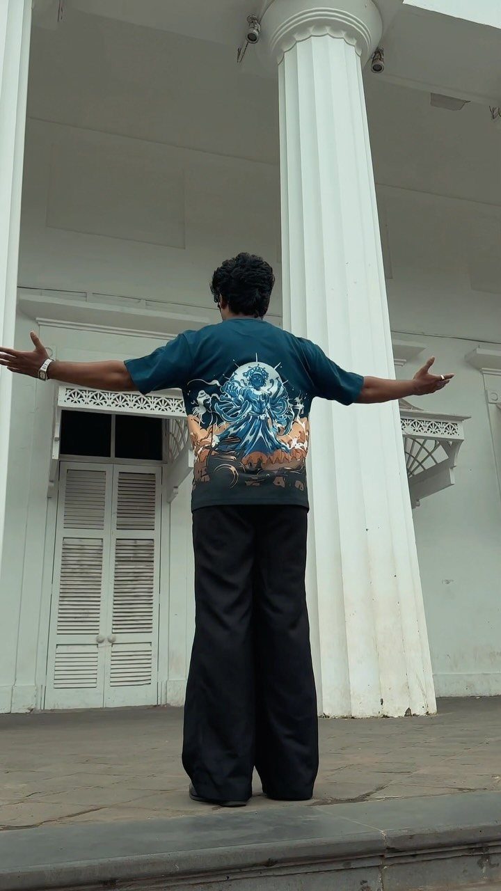 A person wearing a dark teal graphic t-shirt and black wide-leg pants stands before white columns.