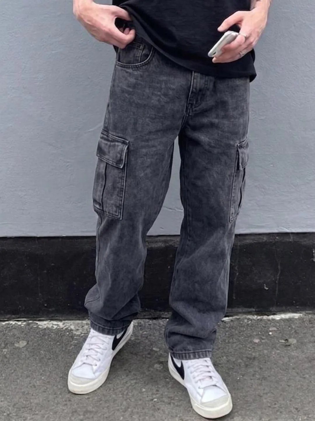 Person wearing dark grey washed cargo jeans and white sneakers standing against a grey wall.
