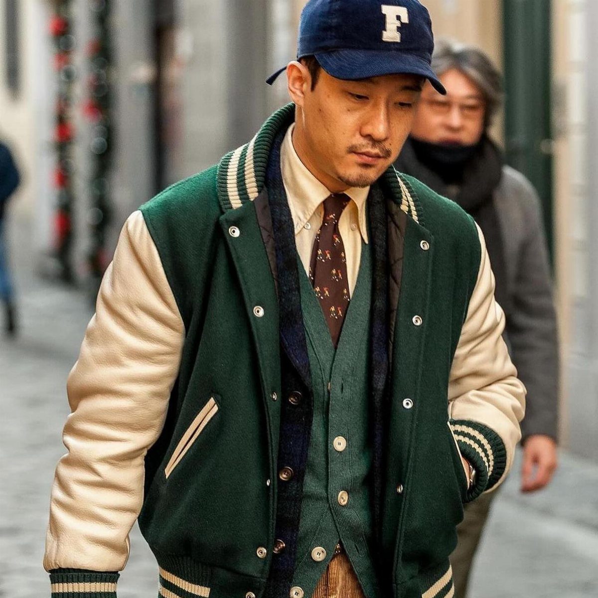 Man wearing a green varsity jacket over a formal shirt, tie, and knit vest.