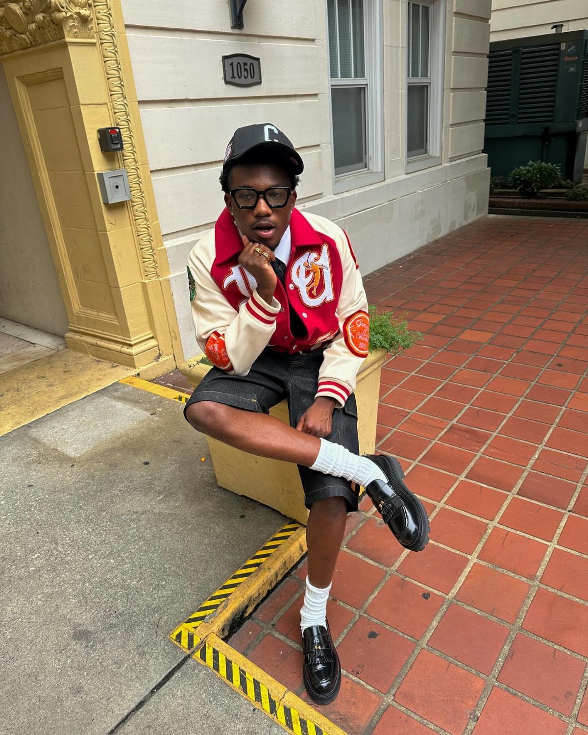 A person in a red and cream varsity jacket and black shorts sitting on a ledge.