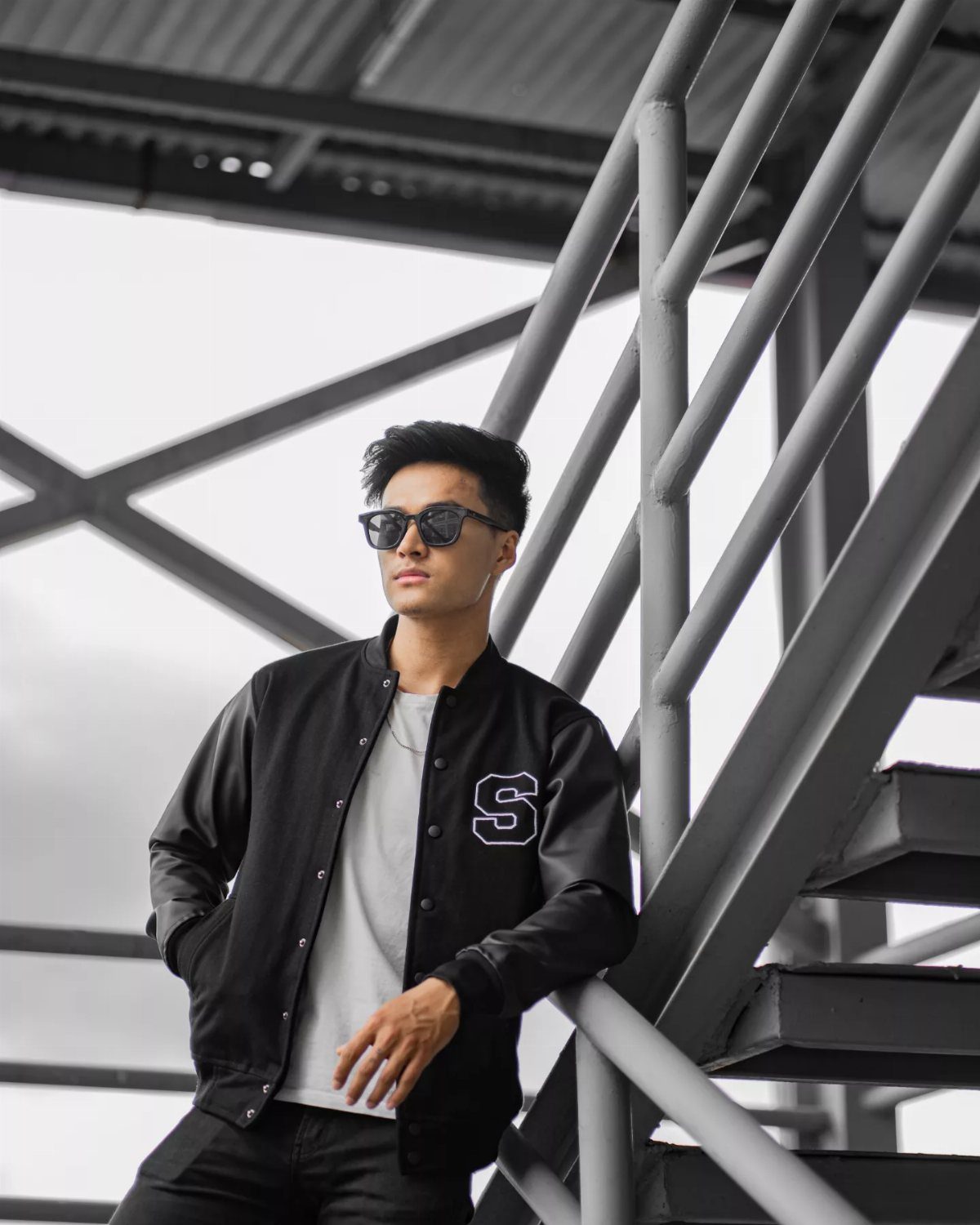 A man in a black varsity jacket and sunglasses leaning against a metal staircase.