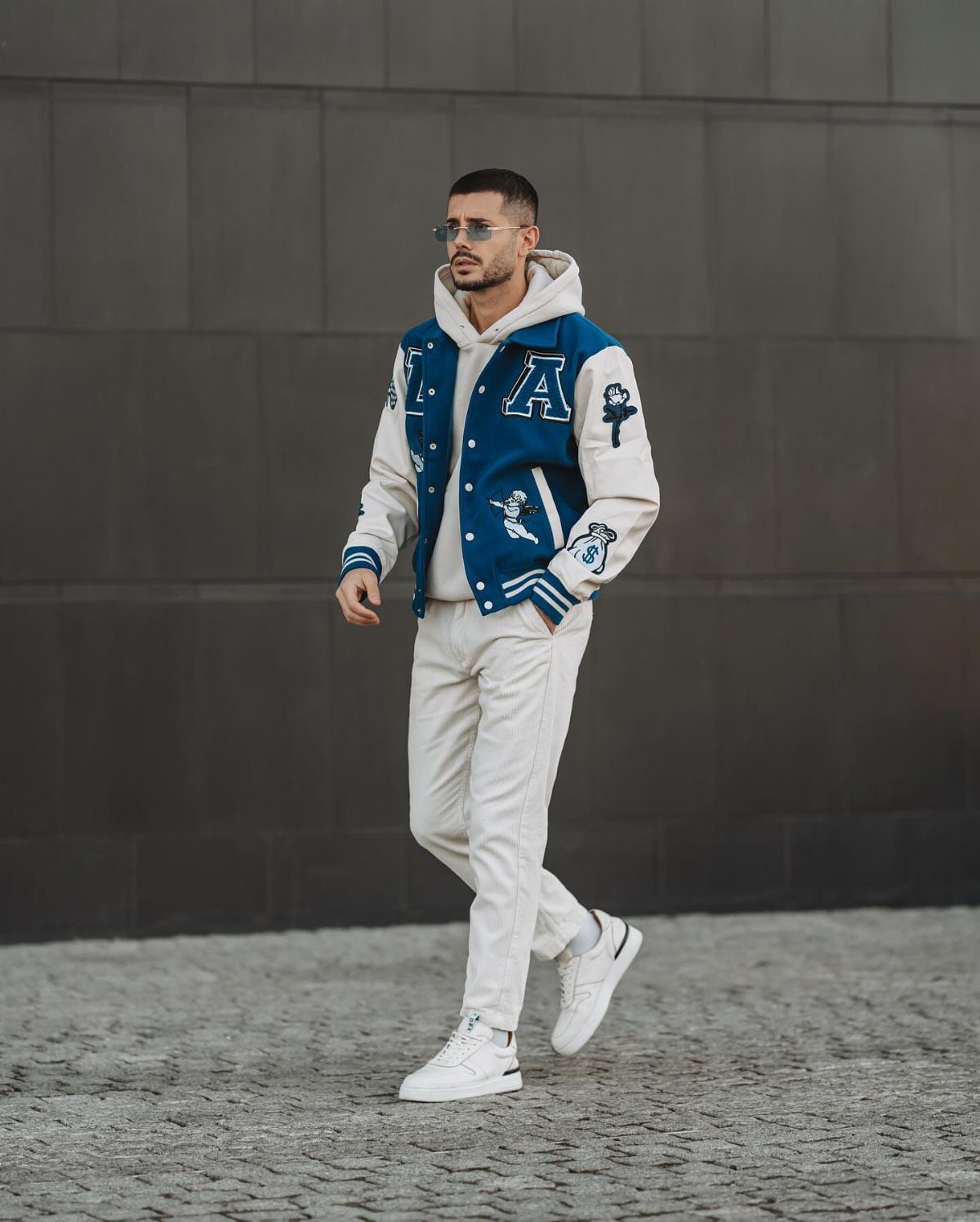 A man walking in a blue and white varsity jacket, white pants, and white sneakers.