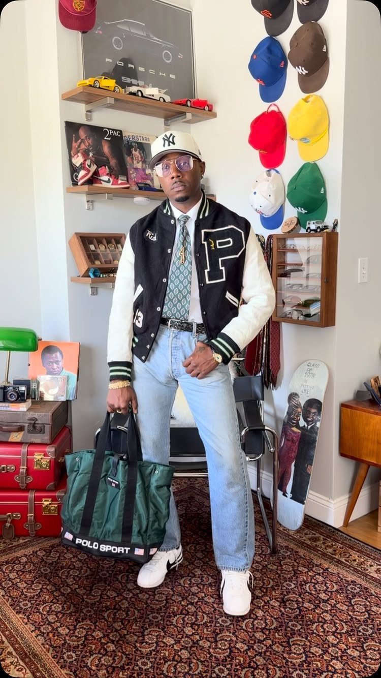 Man in a varsity jacket, light blue jeans, and white sneakers standing in a room.