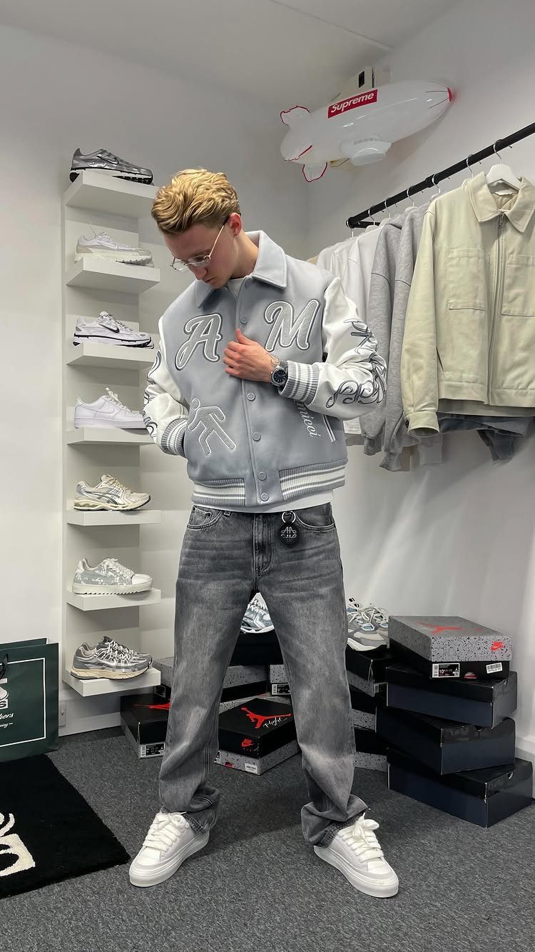 Young man wearing a grey varsity jacket, grey jeans, and white sneakers in a clothing store.