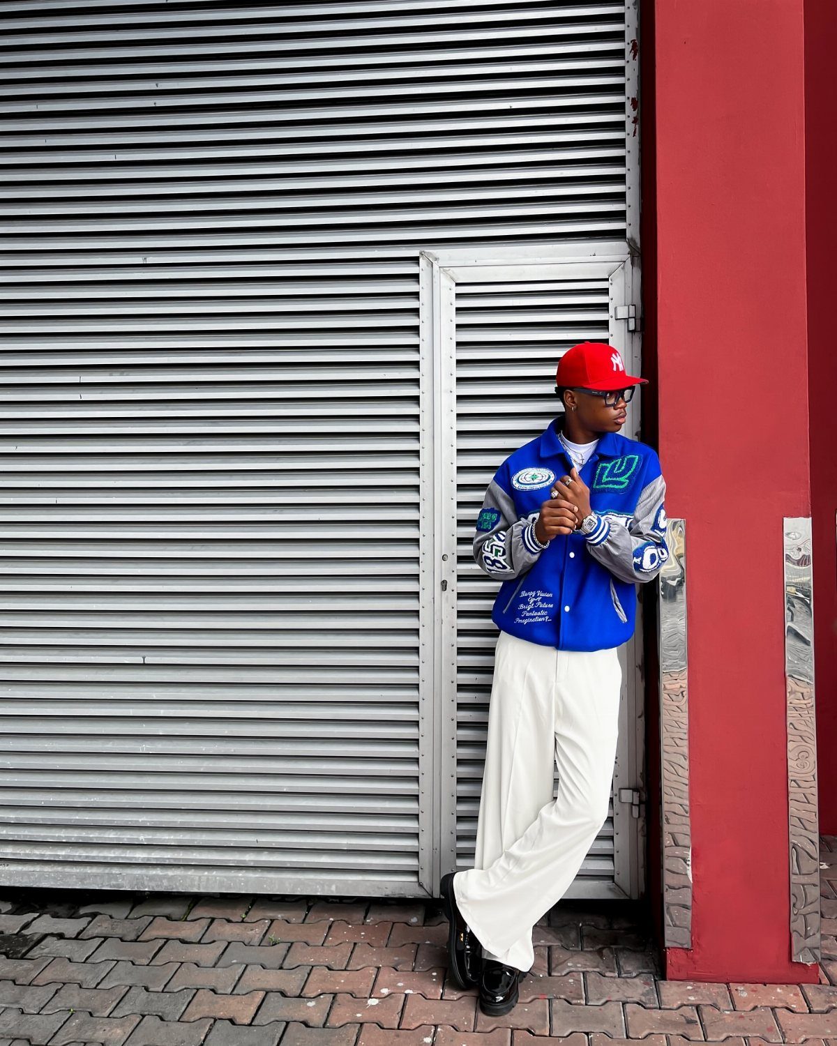 Man in a blue varsity jacket, red cap, and white wide-leg trousers leaning against a metal shutter.