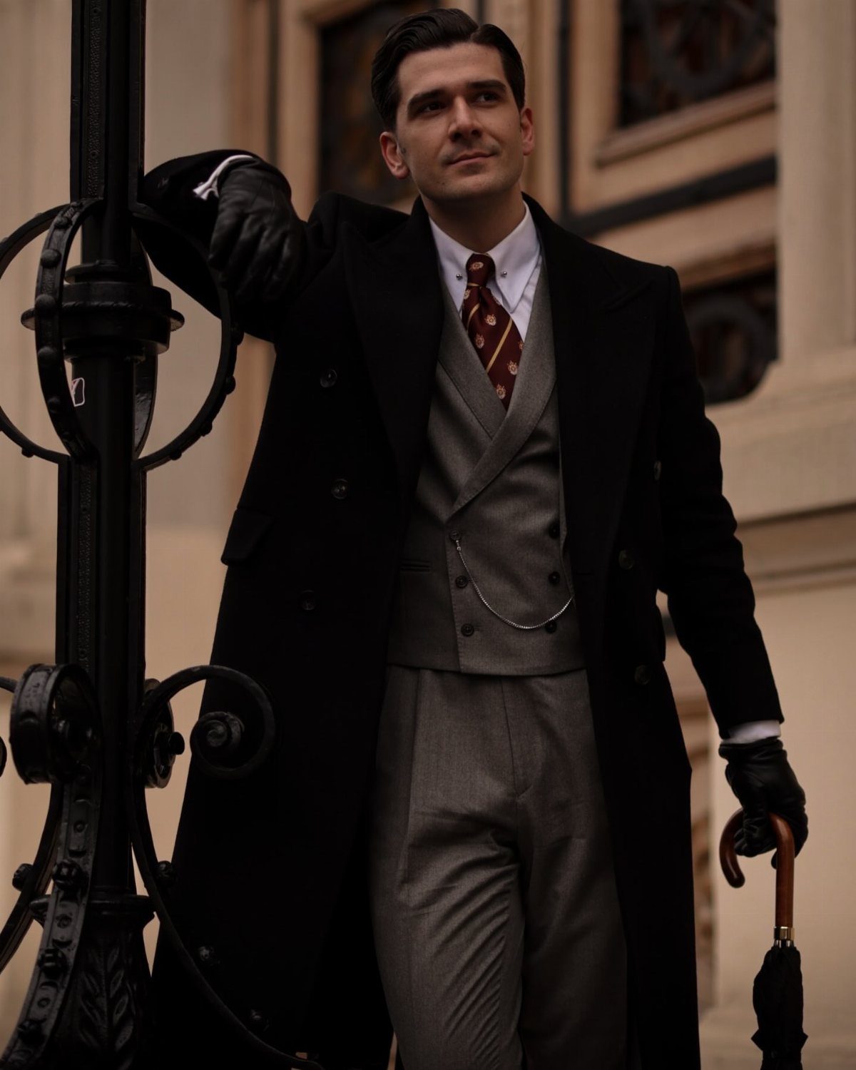 Man in Victorian-style black overcoat and grey waistcoat standing by an ornate metal post.