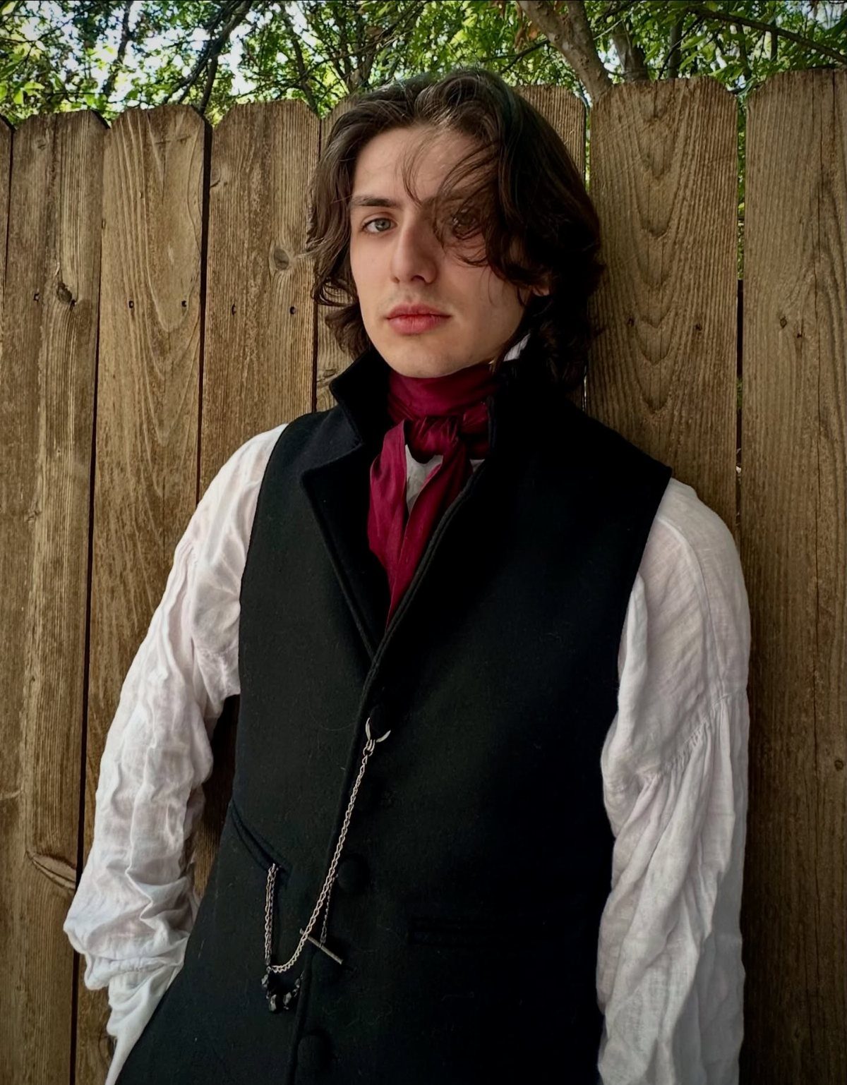 A young man in Victorian-style clothing posing against a wooden fence outdoors.