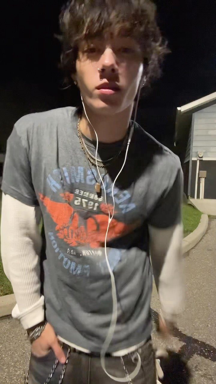 A young person with messy hair wearing a layered vintage graphic t-shirt and necklaces outdoors at night.