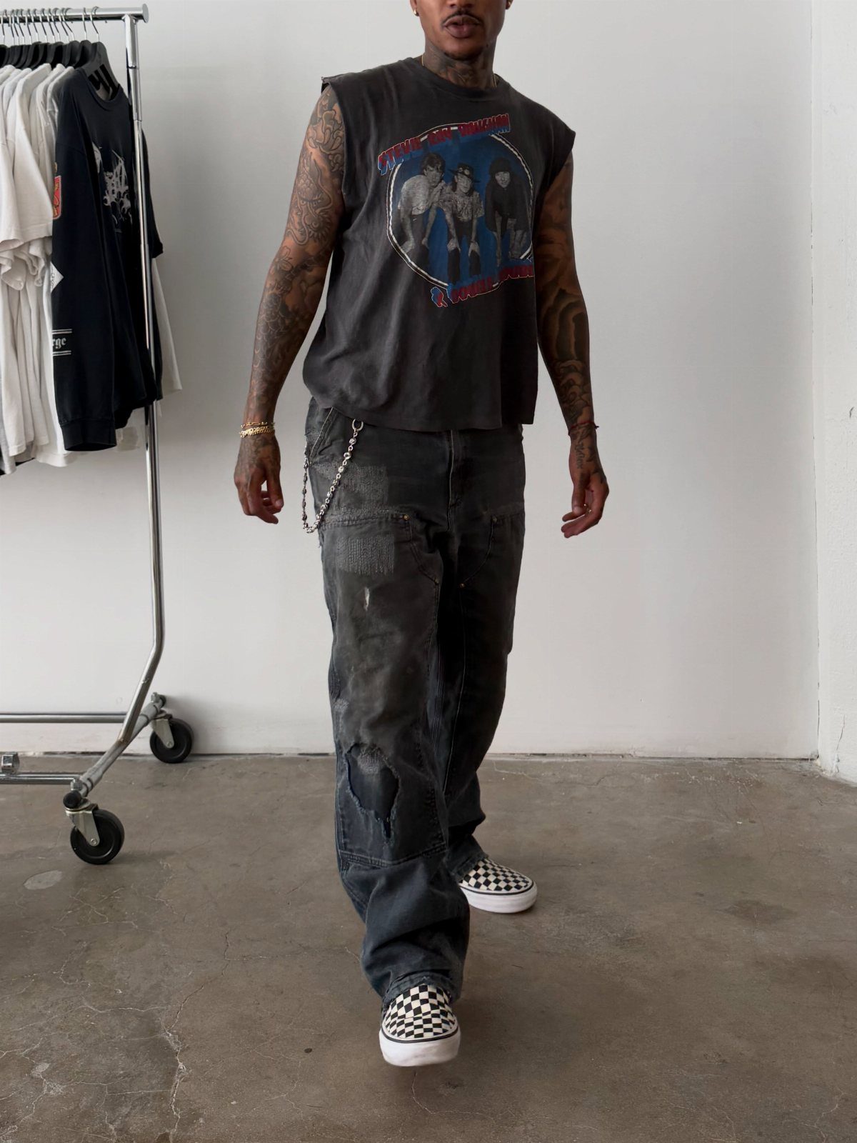 Man wearing a sleeveless graphic tee, baggy distressed jeans, and checkered slip-on sneakers.
