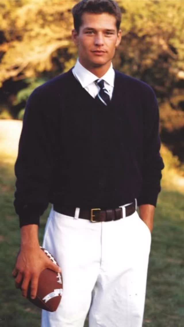 A young man in a navy sweater, white shirt, and tie holding a football outdoors.