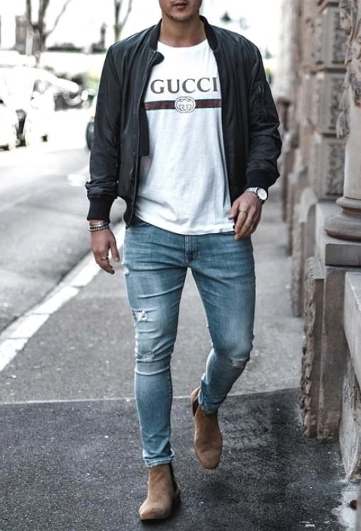 A man walking outdoors wearing a black bomber jacket, white graphic t-shirt, and blue skinny jeans.