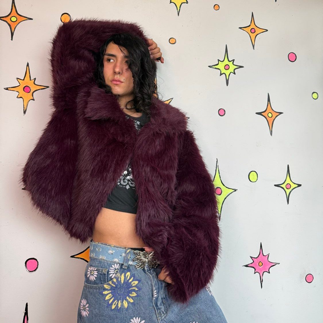 A person wearing a large burgundy faux fur coat and embroidered denim jeans against a star-patterned wall.