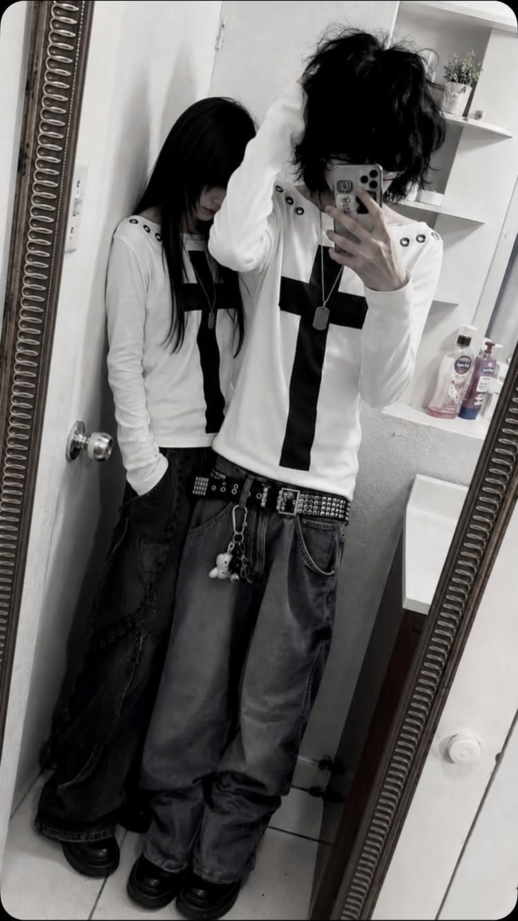 Person in a white cross shirt and baggy grey pants posing for a mirror selfie.