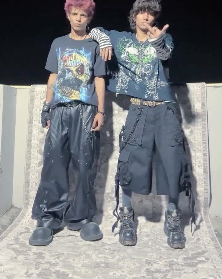 Two people posing in dark graphic tees, faux leather pants, and cargo shorts with heavy boots.